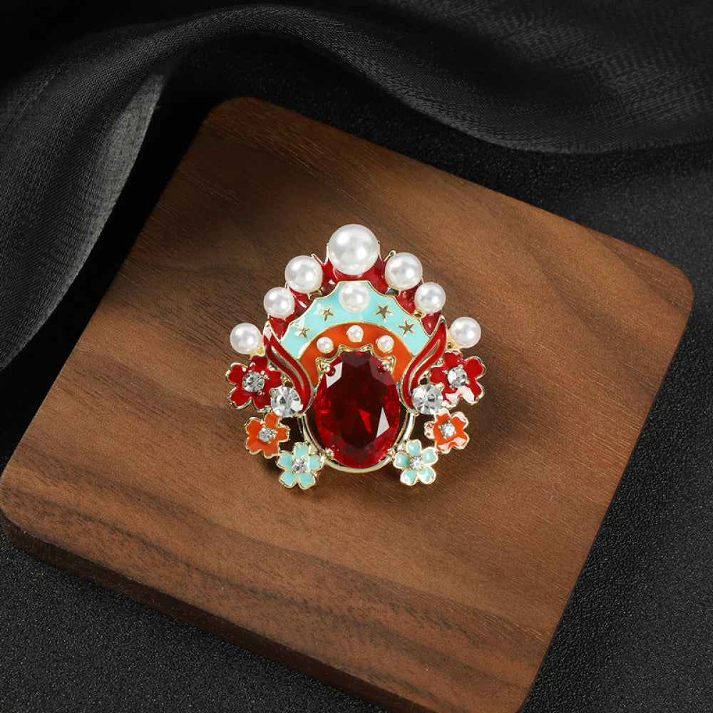 This striking brooch features a bold, theatrical-inspired design with vibrant enamel, lustrous pearls, and a prominent red crystal at its center. Delicate floral motifs and sparkling crystals add intricate detail, making it a standout accessory for blazers, scarves, or dresses. Perfect for adding a pop of color and artistic flair to any outfit, this piece is sure to make a memorable statement.