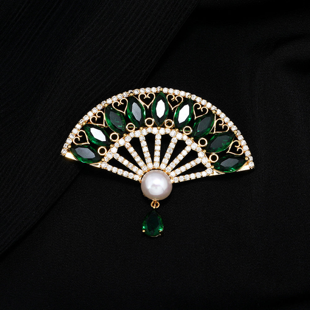This elegant brooch features a striking fan-shaped design, crafted with a gold-tone metal base. It is adorned with vibrant emerald-green gemstones, shimmering clear crystals, and a lustrous pearl at the center, from which a teardrop-shaped green gem dangles gracefully. Perfect for adding a touch of sophistication to blazers, scarves, or evening wear, this piece blends timeless elegance with a bold, eye-catching silhouette.