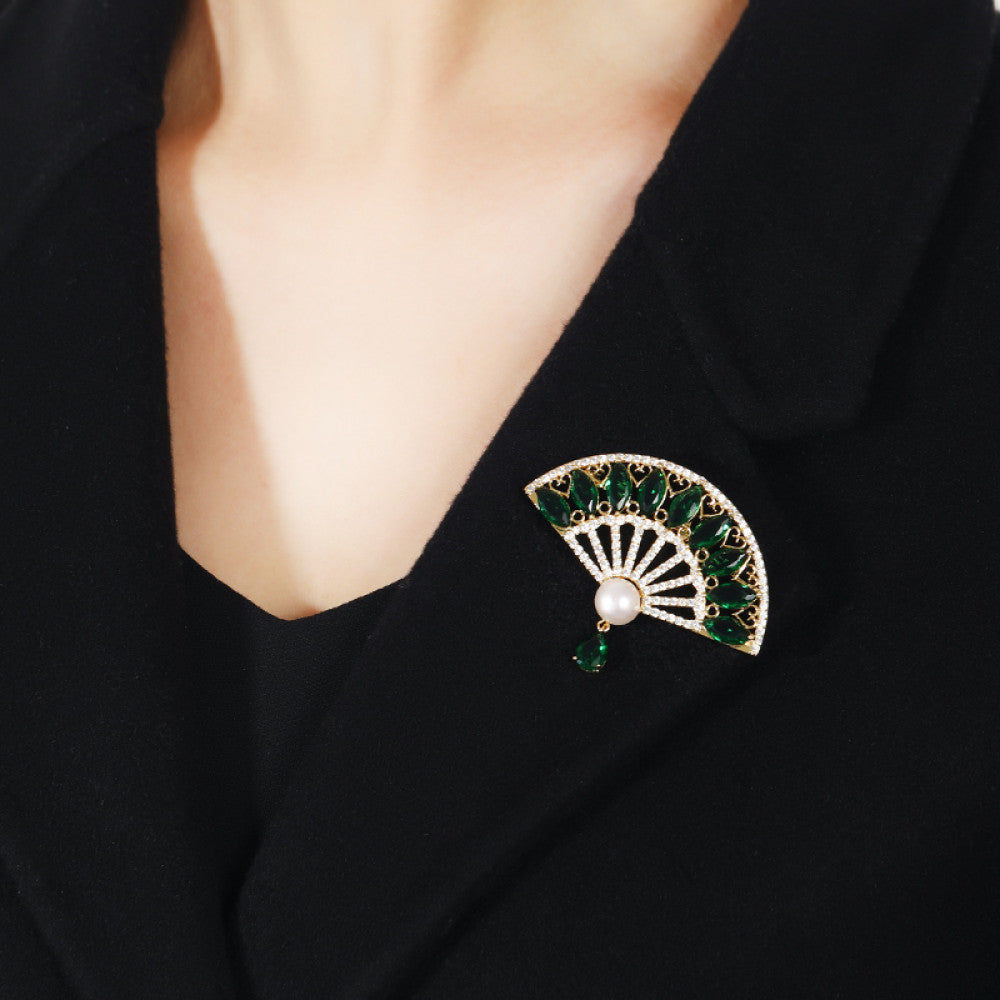 This elegant brooch features a striking fan-shaped design, crafted with a gold-tone metal base. It is adorned with vibrant emerald-green gemstones, shimmering clear crystals, and a lustrous pearl at the center, from which a teardrop-shaped green gem dangles gracefully. Perfect for adding a touch of sophistication to blazers, scarves, or evening wear, this piece blends timeless elegance with a bold, eye-catching silhouette. 01