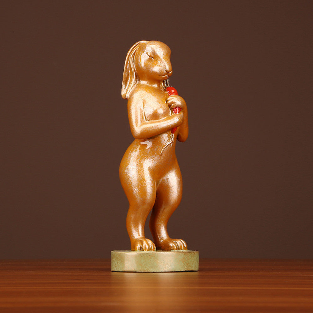 Golden rabbit statue in a standing pose holding a red accent, a symbol of good luck for home decor, placed on a greenish base against a brown background 