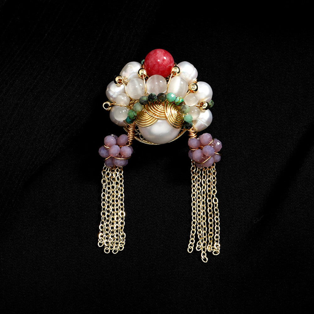 This artisan-crafted brooch features intricate gold wire-wrapping, showcasing a central arrangement of lustrous pearls, a vibrant red gemstone, and rich green accents. Delicate purple bead clusters dangle from either side, cascading into elegant gold tassels that sway with movement. Perfect for adding a touch of bohemian luxury to blazers, scarves, or dresses, this statement piece is a true work of wearable art.