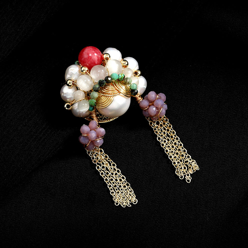 This artisan-crafted brooch features intricate gold wire-wrapping, showcasing a central arrangement of lustrous pearls, a vibrant red gemstone, and rich green accents. Delicate purple bead clusters dangle from either side, cascading into elegant gold tassels that sway with movement. Perfect for adding a touch of bohemian luxury to blazers, scarves, or dresses, this statement piece is a true work of wearable art. 01