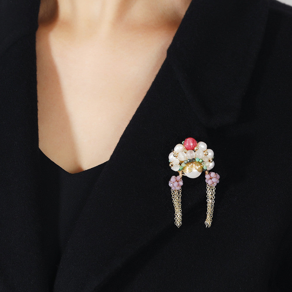 This artisan-crafted brooch features intricate gold wire-wrapping, showcasing a central arrangement of lustrous pearls, a vibrant red gemstone, and rich green accents. Delicate purple bead clusters dangle from either side, cascading into elegant gold tassels that sway with movement. Perfect for adding a touch of bohemian luxury to blazers, scarves, or dresses, this statement piece is a true work of wearable art. 02