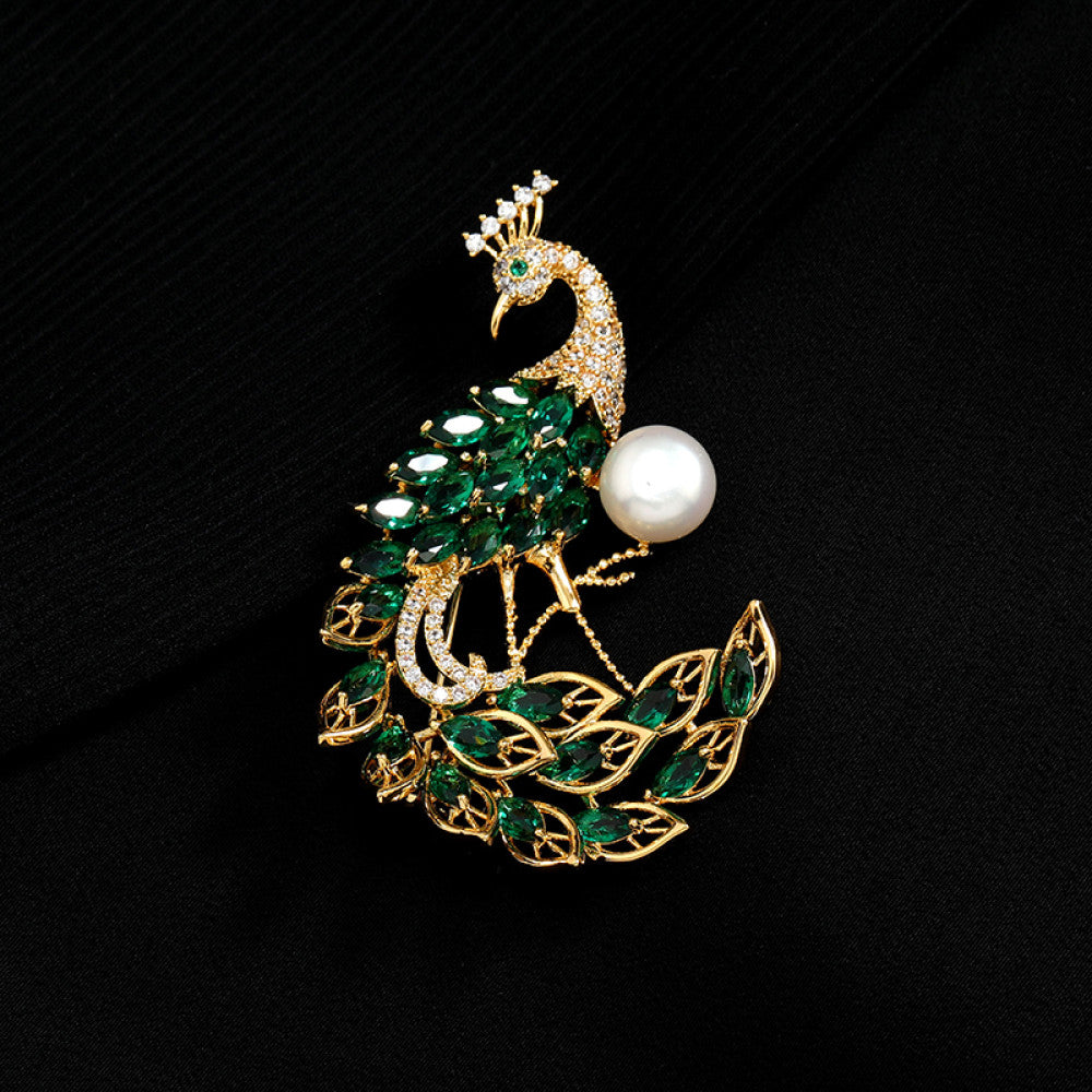 This opulent brooch showcases a majestic peacock design, crafted with a gleaming gold-tone metal base. The peacock’s body and elaborate tail are lavishly adorned with vibrant emerald-cut green crystals, while its head and neck are encrusted with sparkling clear crystals. A large, lustrous white pearl rests gracefully beside the peacock, adding a touch of timeless elegance. Perfect for elevating formal attire, evening gowns, or special occasion outfits, this statement piece exudes luxury and intricate crafts