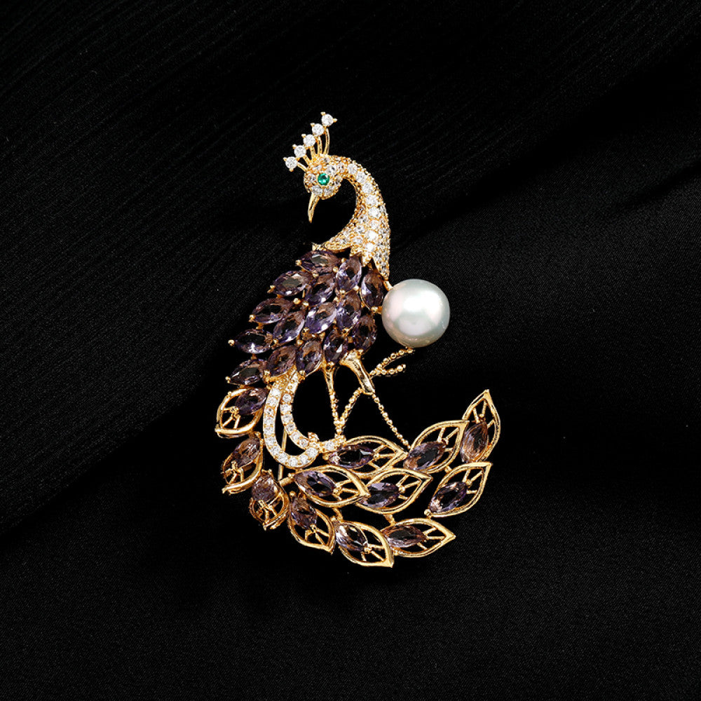 This opulent brooch showcases a majestic peacock design, crafted with a gleaming gold-tone metal base. The peacock’s body and elaborate tail are lavishly adorned with vibrant emerald-cut green crystals, while its head and neck are encrusted with sparkling clear crystals. A large, lustrous white pearl rests gracefully beside the peacock, adding a touch of timeless elegance. Perfect for elevating formal attire, evening gowns, or special occasion outfits