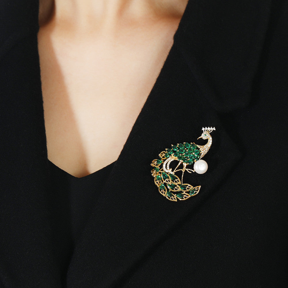 This opulent brooch showcases a majestic peacock design, crafted with a gleaming gold-tone metal base. The peacock’s body and elaborate tail are lavishly adorned with vibrant emerald-cut green crystals, while its head and neck are encrusted with sparkling clear crystals. A large, lustrous white pearl rests gracefully beside the peacock, adding a touch of timeless elegance. Perfect for elevating formal attire, evening gowns, or special occasion outfits 02