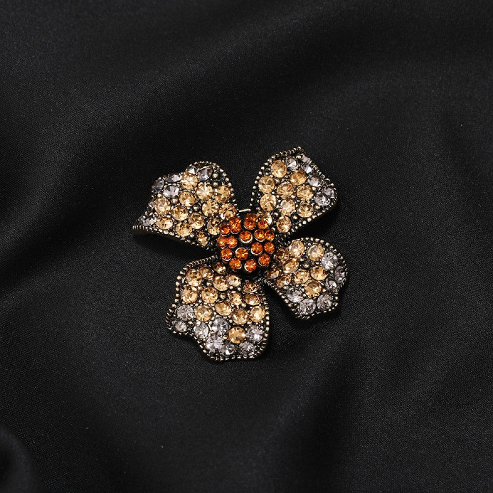 This eye-catching brooch features a striking four-petal floral design, crafted on an antiqued metal base and lavishly adorned with a dazzling array of crystals. Each petal shimmers with a gradient of clear and golden crystals, while a vibrant cluster of orange crystals anchors the center, creating a bold, layered effect. Perfect for adding a touch of vintage glamour to blazers, scarves, or evening wear, this piece blends intricate craftsmanship with timeless, eye-catching charm