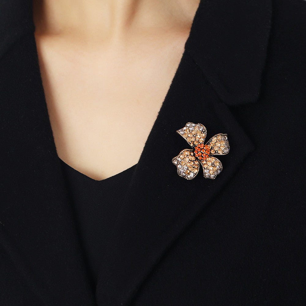 This eye-catching brooch features a striking four-petal floral design, crafted on an antiqued metal base and lavishly adorned with a dazzling array of crystals. Each petal shimmers with a gradient of clear and golden crystals, while a vibrant cluster of orange crystals anchors the center, creating a bold, layered effect. Perfect for adding a touch of vintage glamour to blazers, scarves, or evening wear, this piece blends intricate craftsmanship with timeless, eye-catching charm 01