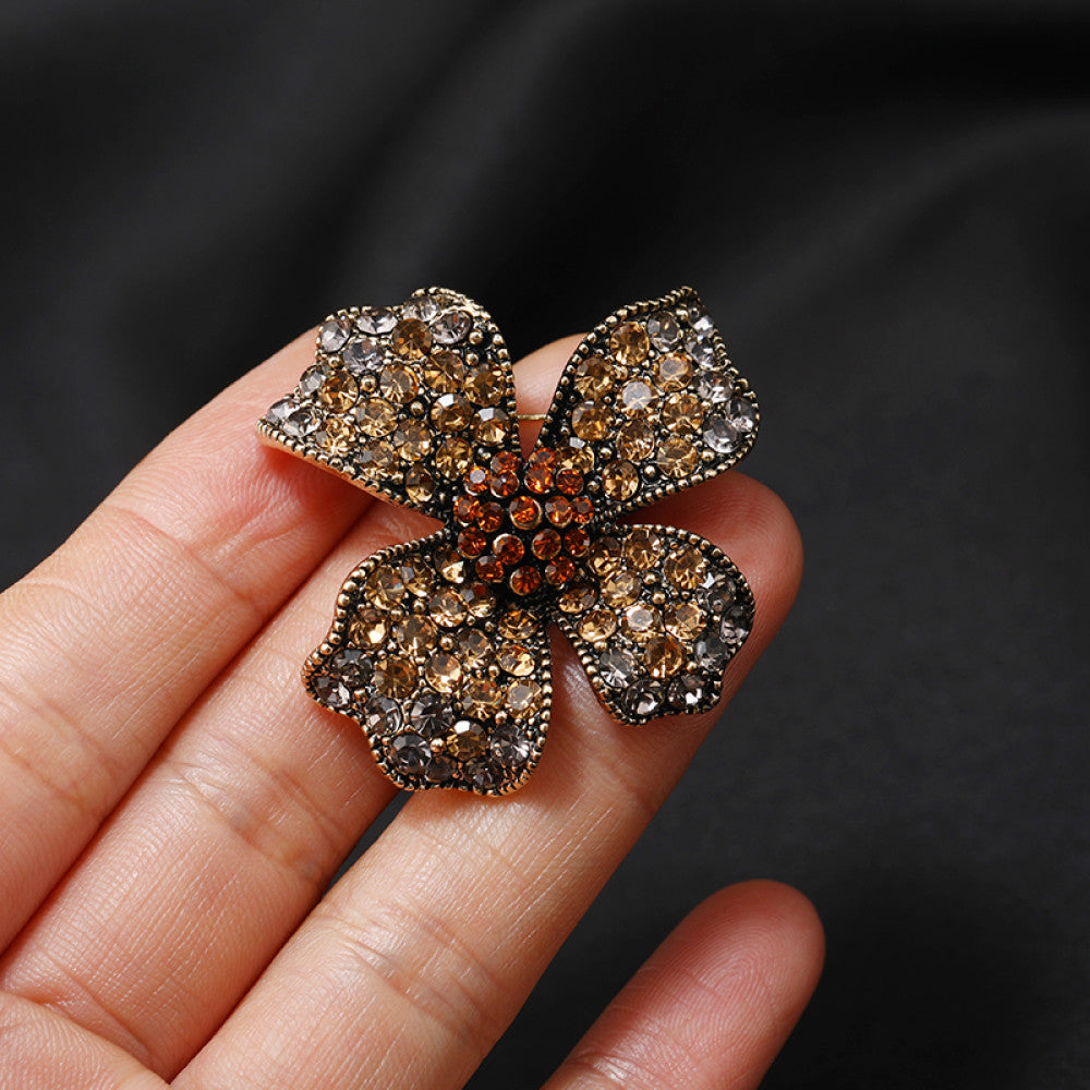 This eye-catching brooch features a striking four-petal floral design, crafted on an antiqued metal base and lavishly adorned with a dazzling array of crystals. Each petal shimmers with a gradient of clear and golden crystals, while a vibrant cluster of orange crystals anchors the center, creating a bold, layered effect. Perfect for adding a touch of vintage glamour to blazers, scarves, or evening wear, this piece blends intricate craftsmanship with timeless, eye-catching charm 02
