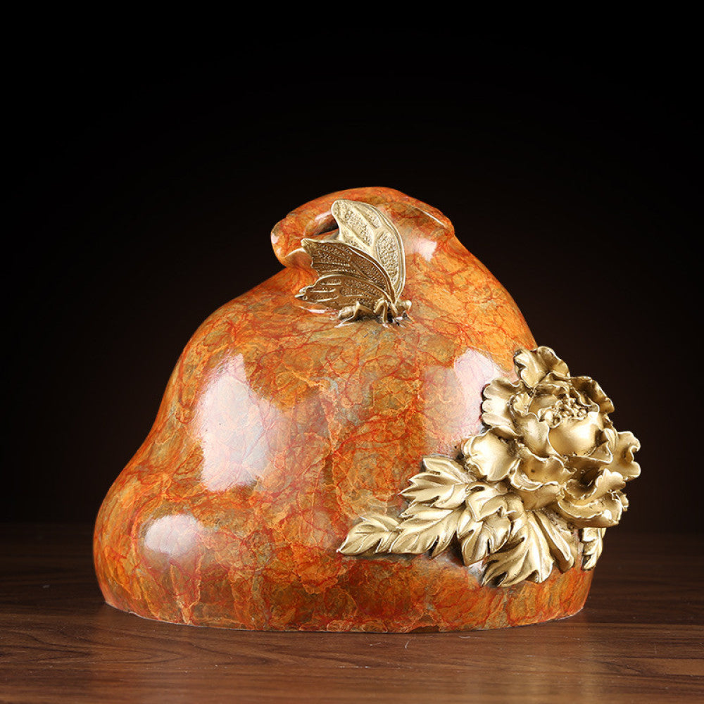 Orange textured decorative sculpture with brass peony flower and butterfly details, displayed on wooden table against dark background 
