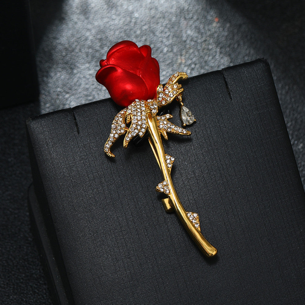 This elegant brooch features a vivid red rose with a gold crystal-encrusted stem, accented by a delicate teardrop detail. A romantic accessory to elevate any outfit
