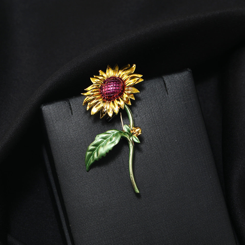 This charming brooch captures the warmth of a sunflower, with intricately detailed gold petals radiating from a rich purple center. Delicate green enamel leaves add a touch of natural vibrancy, while the slender gold stem completes the lifelike design. Perfect for adding a cheerful, nature-inspired touch to blazers, scarves, or everyday outfits, this piece blends whimsical charm with timeless elegance 01