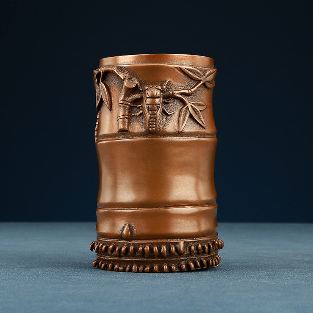 Red copper brush pot with bamboo carvings, holding pens, placed on wooden table against brown background 01