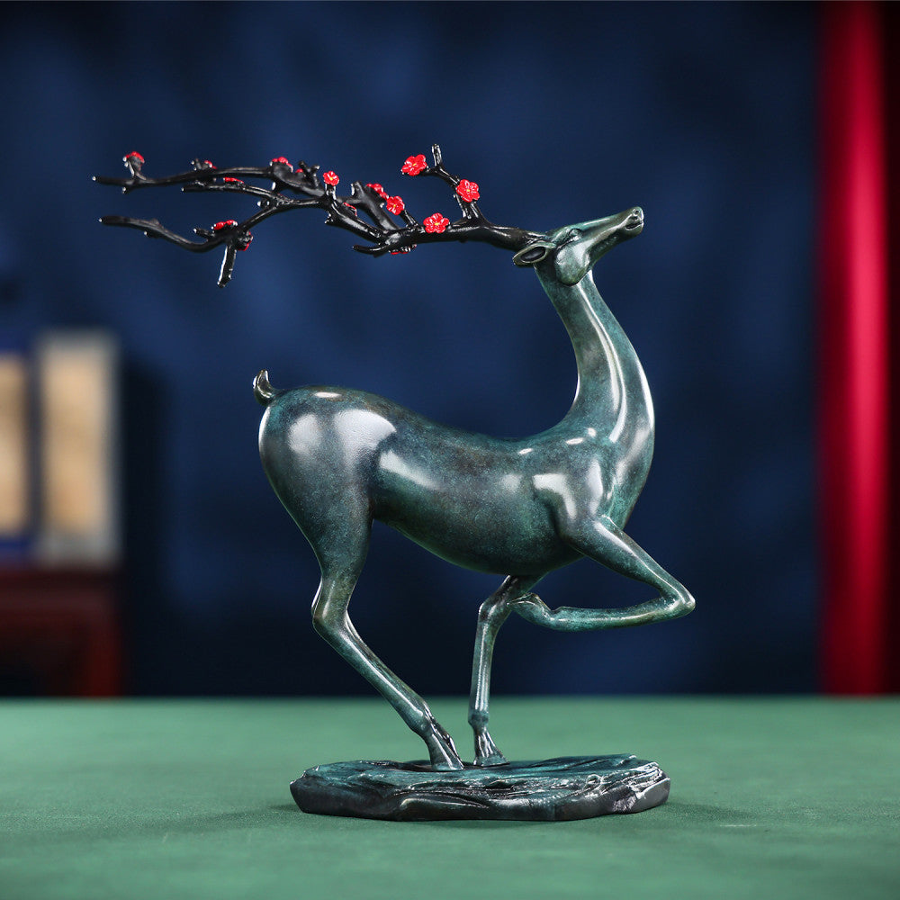 Traditional Chinese bronze deer statue with red plum blossom branch, in an elegant pose, placed on a green surface against a dark blue background