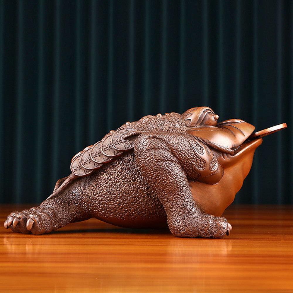 Traditional Chinese bronze money toad statue with a copper coin in its mouth, positioned on a wooden surface against a dark curtain background