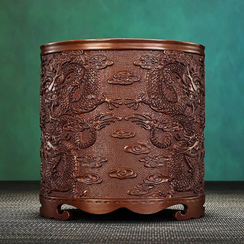 Red copper brush pot with intricate dragon and cloud carvings, placed on a textured surface against a teal background