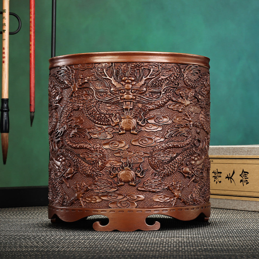 Red copper brush pot with intricate dragon and cloud carvings, placed on a textured surface against a teal background 01