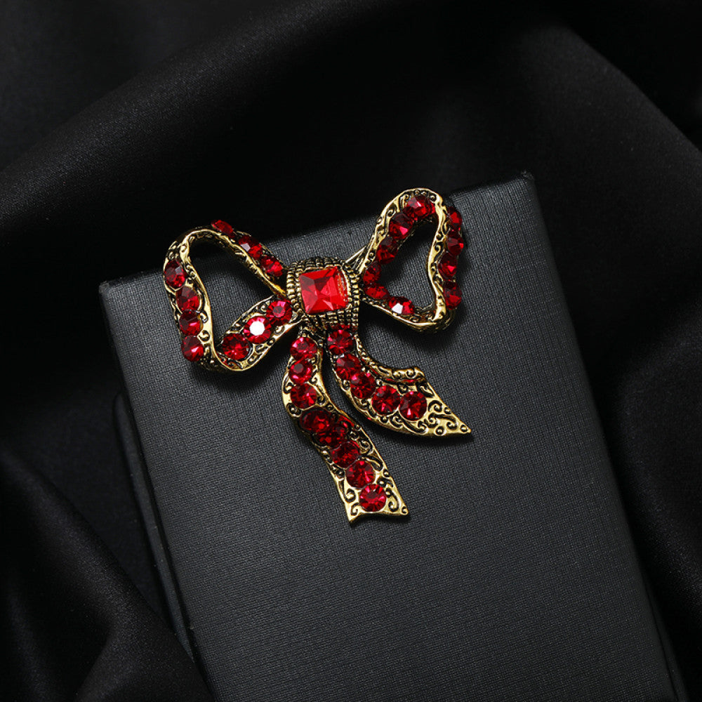 This eye-catching brooch features a classic bow design, crafted with an antiqued gold-tone metal base intricately adorned with vibrant red crystals. A large, faceted red gemstone anchors the center, while delicate filigree detailing adds vintage charm. Perfect for elevating blazers, scarves, or festive outfits, this bold piece brings a touch of glamorous retro style to any look.
