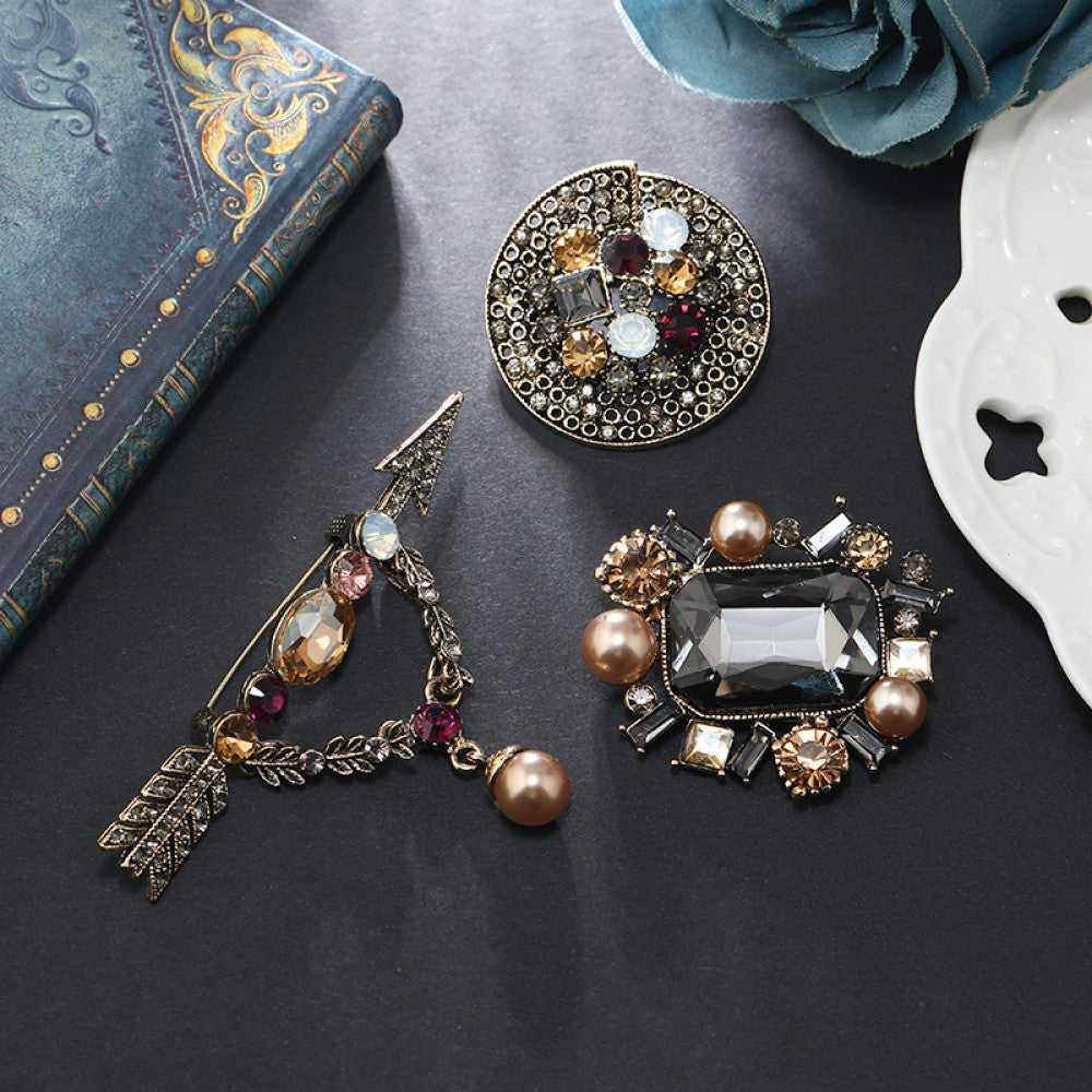 This vintage brooch set features an antiqued bronze base, adorned with pearls, colored crystals, and gemstones. The circular design sparkles with dense small crystals; the arrow-shaped piece carries a tribal vibe with a pearl pendant; the large gemstone brooch boasts a bold black stone with surrounding pearls and gems. Ideal for elevating the sophistication and vintage charm of your coats, blazers, or dresses. 