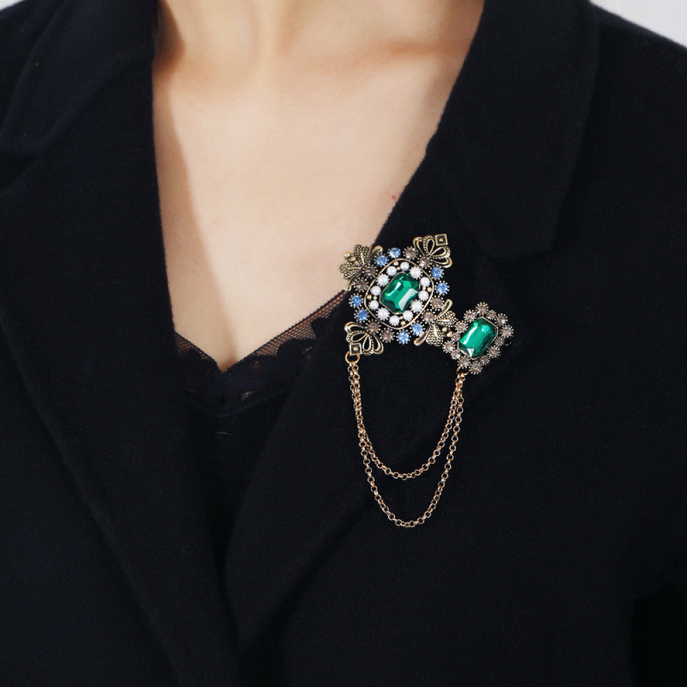 This vintage-style double brooch set features an ornate filigree design with a large emerald-green gem at its center, connected by delicate chains to a smaller matching brooch. Adorned with blue and white accents, it adds sophisticated antique charm to any outfit 02