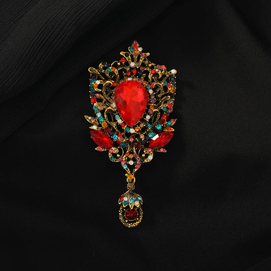 This eye-catching brooch features an ornate, vintage-inspired design with an antiqued gold filigree base. At its center, a large, faceted red crystal takes the spotlight, surrounded by a vibrant array of multicolored crystals and beads. A delicate dangle pendant, adorned with additional gemstones, hangs from the bottom, adding movement and drama. Perfect for elevating blazers, evening gowns, or special occasion outfits, this bold piece exudes opulent, baroque-inspired charm.