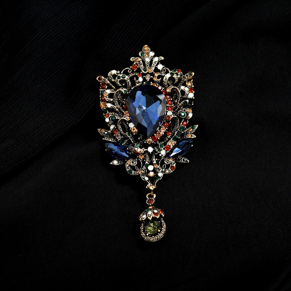 This eye-catching brooch features an ornate, vintage-inspired design with an antiqued gold filigree base. At its center, a large, faceted red crystal takes the spotlight, surrounded by a vibrant array of multicolored crystals and beads. A delicate dangle pendant, adorned with additional gemstones, hangs from the bottom, adding movement and drama. Perfect for elevating blazers, evening gowns, or special occasion outfits, this bold piece exudes opulent, baroque-inspired charm. 01