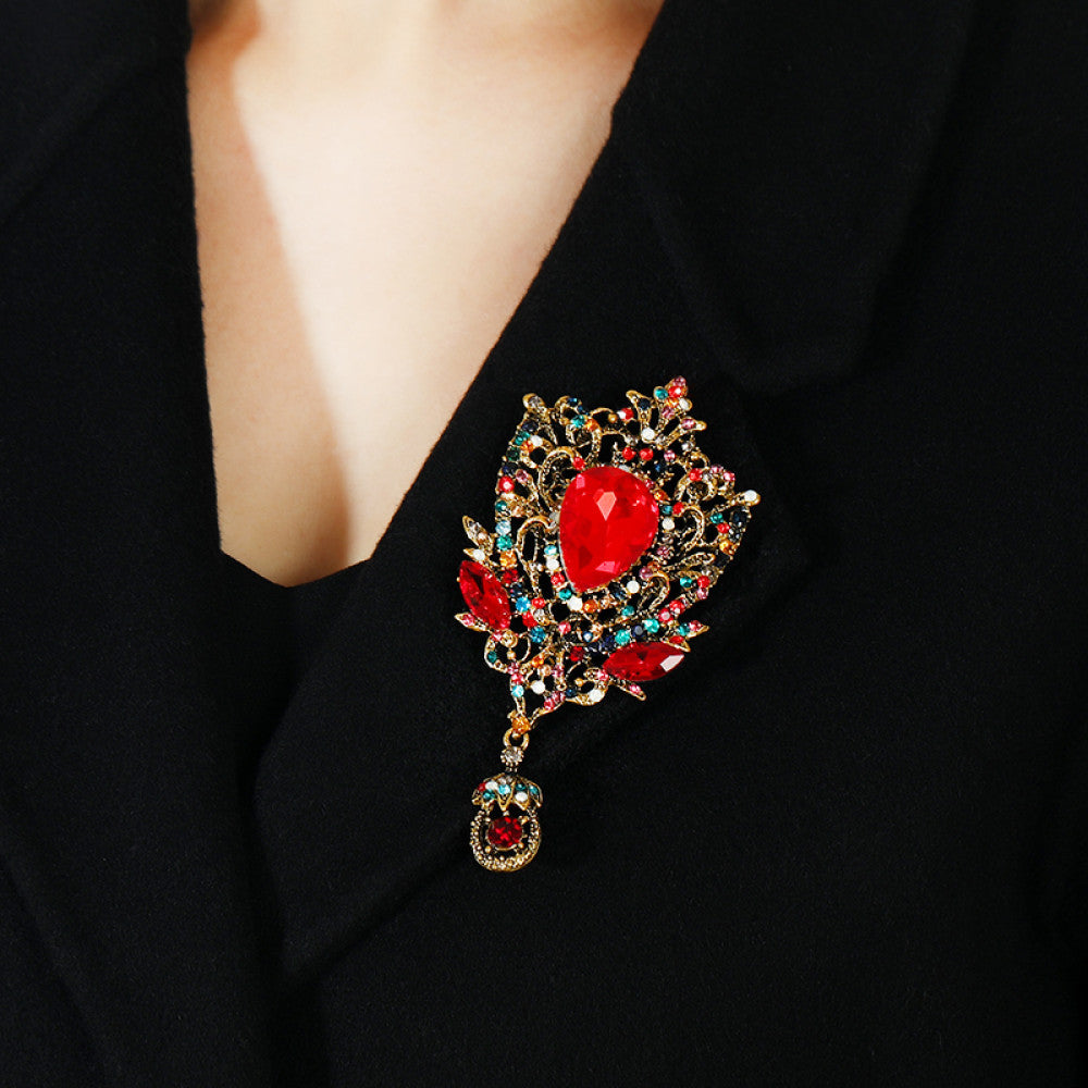 This eye-catching brooch features an ornate, vintage-inspired design with an antiqued gold filigree base. At its center, a large, faceted red crystal takes the spotlight, surrounded by a vibrant array of multicolored crystals and beads. A delicate dangle pendant, adorned with additional gemstones, hangs from the bottom, adding movement and drama. Perfect for elevating blazers, evening gowns, or special occasion outfits, this bold piece exudes opulent, baroque-inspired charm. 03