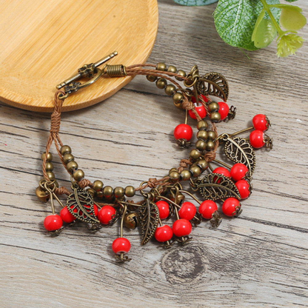 This vintage-style bracelet features a brown braided cord, adorned with antique bronze leaf charms and vibrant red berry-like beads, creating a nature-inspired, bohemian look. The toggle clasp ensures a secure fit, making it a striking accessory for any outfit