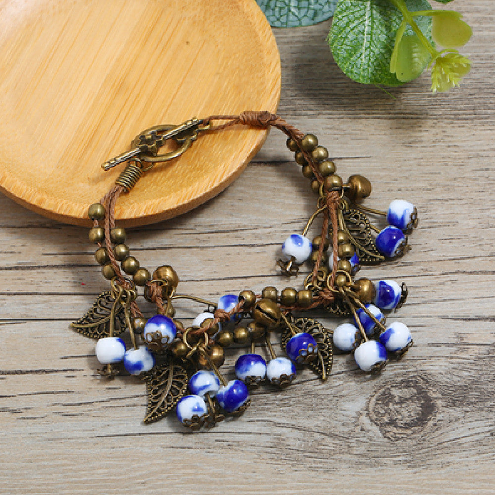 This vintage-style bracelet features a brown braided cord, adorned with antique bronze leaf charms and vibrant red berry-like beads, creating a nature-inspired, bohemian look. The toggle clasp ensures a secure fit, making it a striking accessory for any outfit 1