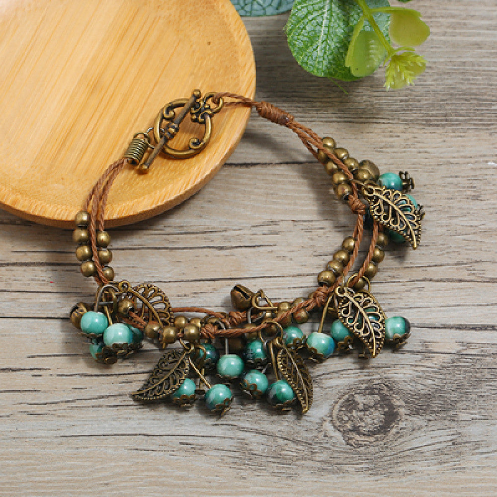 This vintage-style bracelet features a brown braided cord, adorned with antique bronze leaf charms and vibrant red berry-like beads, creating a nature-inspired, bohemian look. The toggle clasp ensures a secure fit, making it a striking accessory for any outfit 2