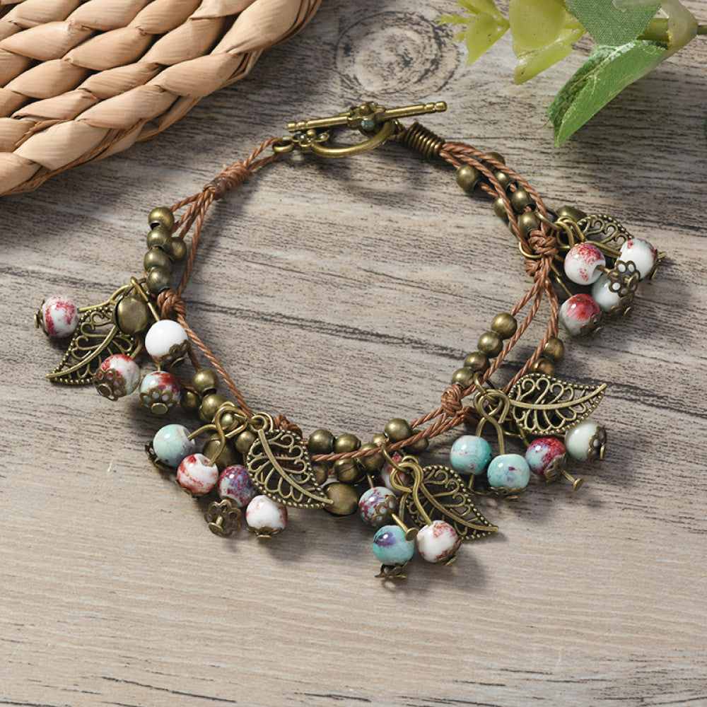 This vintage-style bracelet features a brown braided cord, adorned with antique bronze leaf charms and vibrant red berry-like beads, creating a nature-inspired, bohemian look. The toggle clasp ensures a secure fit, making it a striking accessory for any outfit 7